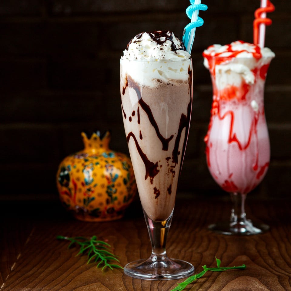Two milkshakes, chocolate and strawberry, topped with whipped cream and syrup, on a wooden table.